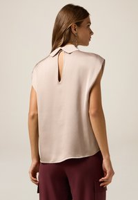 Top senza maniche beige chiaro con colletto, scollatura a chiave sul retro e texture liscia. Abbinato a pantaloni marrone scuro con tasche.