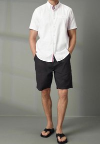 Camicia bianca a maniche corte con bottoni e una sottile trama, abbinata a pantaloni corti neri e infradito neri. Design del colletto interno a contrasto visibile.