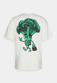 White t-shirt featuring a large, colorful illustration of a cartoon broccoli character with expressive eyes, holding a bag labeled "des légumes."