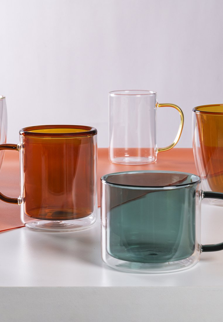 Zestaw szklanek w różnych kształtach i kolorach, w tym bursztynowym, przezroczystym i tealowym. Posiadają gładkie powierzchnie i kontrastujące uchwyty.