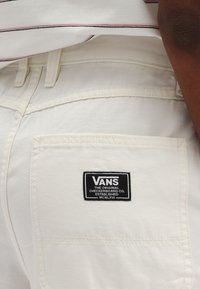 White denim pants featuring a back pocket with a black Vans logo patch. The stitching is visible, adding texture to the fabric.