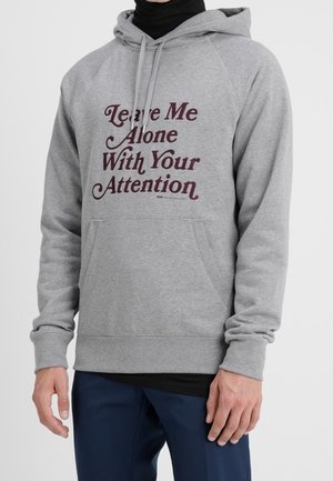 Sudadera con capucha gris con bolsillo tipo canguro, con texto en color burdeos: "Déjame en paz con tu atención." Detalles incluyen capucha con cordón y puños acanalados.