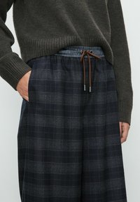 Marineblauwe geruite broek met een elastische tailleband, voorzien van een bruine trekkoord en zijzakken. Gemaakt van een zachte, gestructureerde stof.