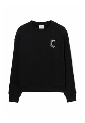 Mujer con cabello largo y oscuro vistiendo una sudadera negra con un logo blanco y pantalones anchos azul con patrón, de pie frente a un fondo liso.