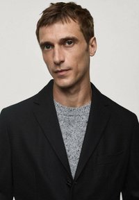 Blazer negru cu un singur nasture, asortat cu un pulover gri cu modele. Materialele par netede, cu linii curate și o croială modernă.