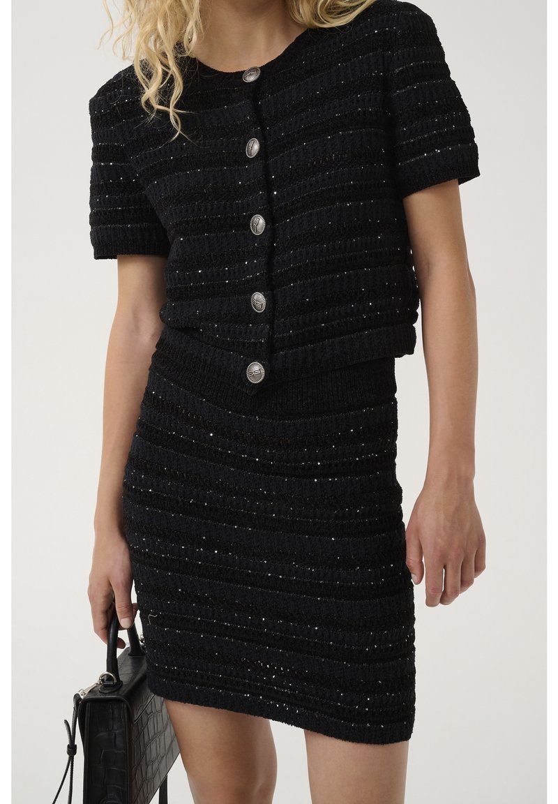 Femme portant un haut à manches courtes texturé noir boutonné et une jupe assortie, tenant un sac à main noir avec un motif crocodile.