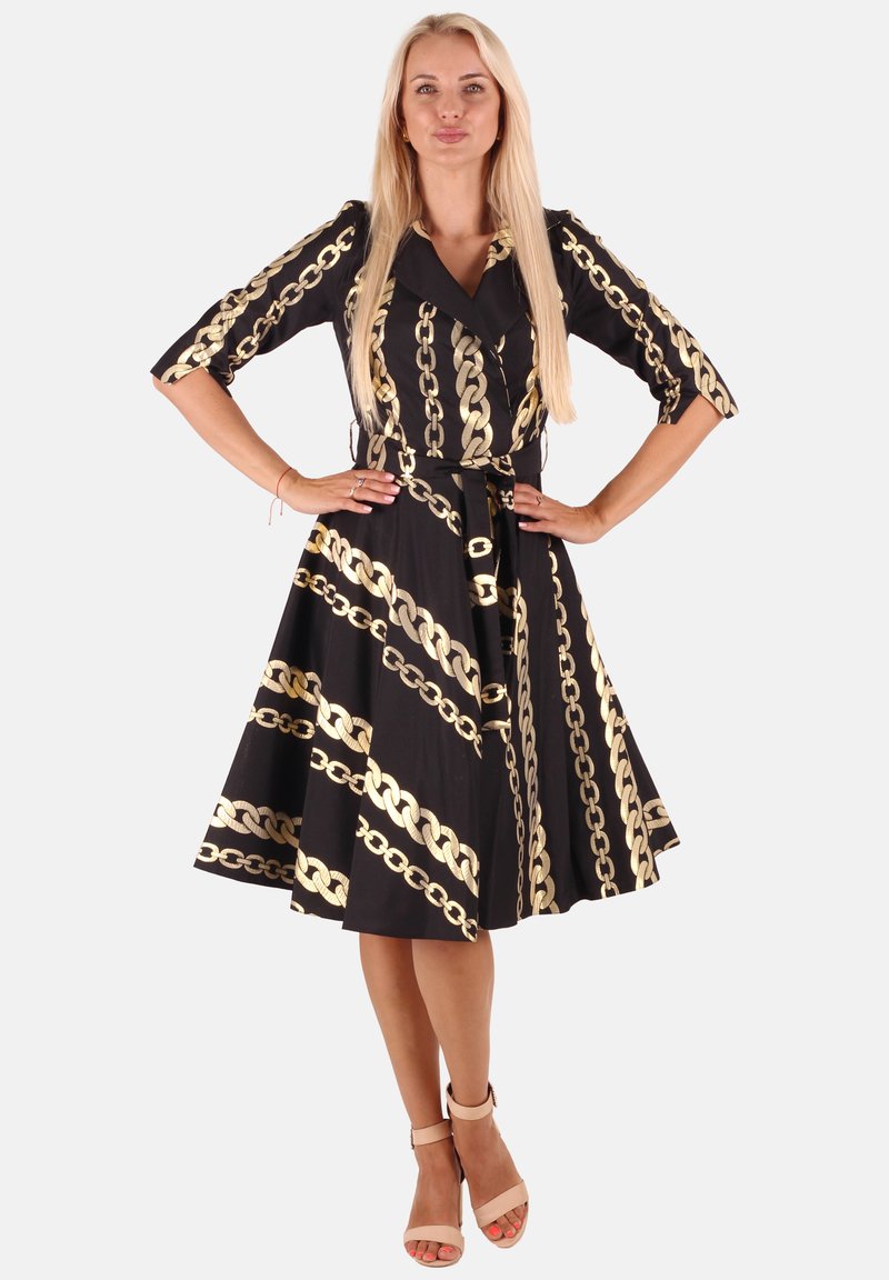 Frau mit langen blonden Haaren, die ein schwarzes knielanges Kleid mit goldenen Kettenmustern trägt, steht mit den Händen auf den Hüften und beigen High-Heels.