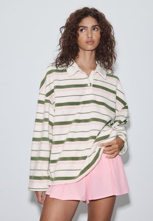 Kvinde med krøllet hår iført en hvid langærmet striped skjorte i grøn og pink, holder kanten med den ene hånd, kombineret med pink shorts.