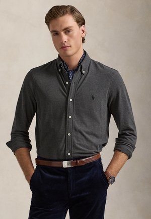 Jeune homme portant une chemise gris foncé à boutons, une écharpe bleu marine à pois, une ceinture en cuir marron, un pantalon bleu marine et une montre-bracelet, debout les mains dans les poches.