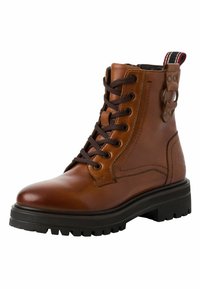 Bota de tobillo de cuero marrón con cordones y suela robusta, con acentos metálicos y una pestaña de tira a rayas en el talón. Textura suave con un acabado pulido.