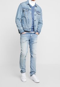 Ljusblå denimjacka, blå tröja, ljusblå jeans och vita sneakers. Denimen har klassiska fickor och sömnadsdetaljer.