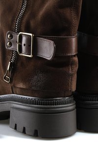 Gros plan de bottes en daim marron foncé avec semelles épaisses en caoutchouc noir, fermeture éclair latérale et sangle en cuir réglable avec boucle métallique.