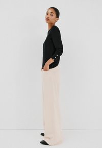 Top nero a maniche lunghe con dettagli a bottoni sulle maniche, abbinato a pantaloni a gamba larga color crema e scarpe basse nere, su uno sfondo neutro.