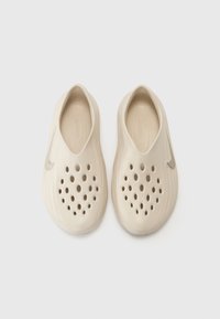 Beige slip-on shoes with multiple ventilation holes, smooth texture, and a silver Nike logo accent on the side. Rounded shape.