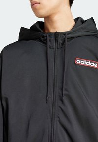 Svart zip-up hoodie med justerbar huva, med en röd broderad "adidas"-logga på vänster bröst och en slät, matt yta.