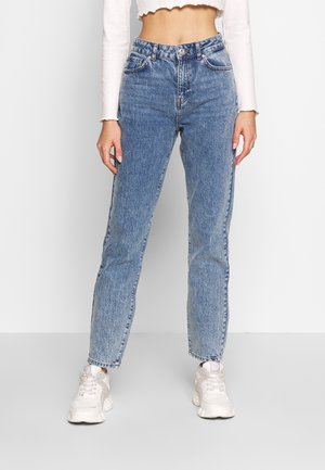 Person iført højttaljede blå jeans og hvide store sneakers står mod en ensartet lys baggrund.