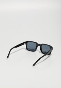 Occhiali da sole neri a forma quadrata con lenti grigio scuro, montatura spessa in plastica e aste nere eleganti con dettagli in metallo argentato.