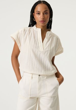 Weiße Kurzarmbluse mit V-Ausschnitt und strukturierten vertikalen Streifen. Kombiniert mit hoch Taillierten, cremefarbenen Hosen mit seitlichen Taschen.