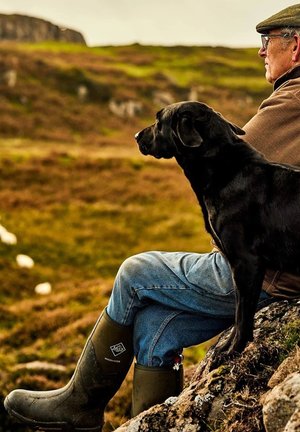 Black labrador sitting closely next to a person wearing green rubber boots and denim jeans, against a natural landscape of grass and rocks.