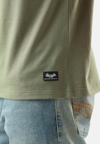Gros plan sur un t-shirt olive vert foncé avec un patch noir montrant un œil ailé et "California 93060 USA" sur un jeans en denim bleu avec des coutures oranges.