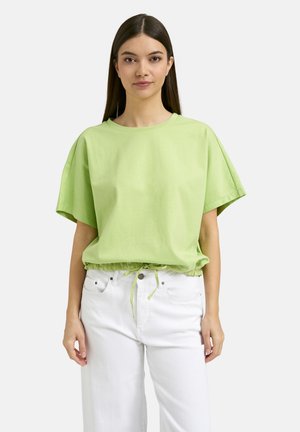 Woman with straight dark hair wearing a light green short-sleeve top with drawstring waist and white pants standing against a plain background.