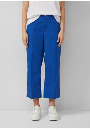 Personne portant un pantalon court bleu vif avec des revers, une chemise blanche rentrée et des baskets blanches sur un fond gris uni.
