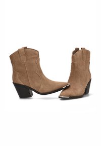 Botas de ante de tobillo en color marrón con punteras puntiagudas, costuras decorativas y un tacón grueso. Presenta acentos plateados en las punteras y en la parte trasera.