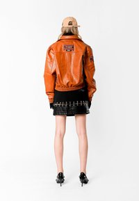 Chaqueta bomber de cuero marrón con puños y dobladillo acanalados, combinada con una falda corta de cuero negro con acentos de metal.