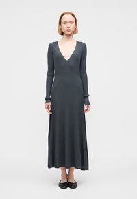 Μακρύ, σκούρο γκρι ριγωτό φόρεμα με βαθύ V ντεκολτέ, μακριά μανίκια με τρύπες για τους αντίχειρες και φαρδύ φούστα με κυματιστή τελείωμα. Φαίνονται επίπεδα παπούτσια.