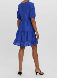 Královsky modré šaty s nafouknutými rukávy a spodní částí s vrstvenými volány, texturovaná tkanina a detaily v oblasti zad se štěrbinou. Nosené s černými sandály na podpatku.