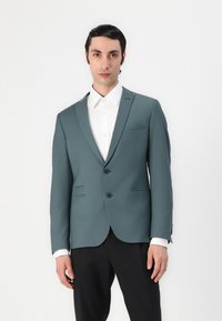 Blazer aderente teal con due bottoni, rever a scendere e pantaloni neri. Presenta imbottiture sulle spalle e due tasche frontali. Camicia bianca sotto.
