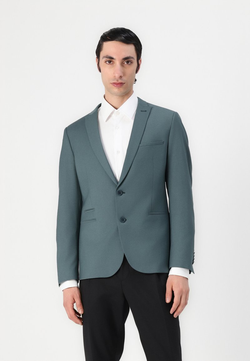 Blazer aderente teal con due bottoni, rever a scendere e pantaloni neri. Presenta imbottiture sulle spalle e due tasche frontali. Camicia bianca sotto.