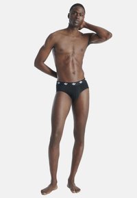 Zwarte katoenen boxershorts met een elastische tailleband met drie witte Adidas-logo's. Het model staat met één hand achter zijn hoofd en de voeten uit elkaar.