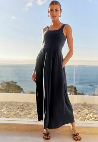 Frau in einem ärmellosen, marineblauen Maxikleid mit Taschen und schwarzen Sandalen, die auf einer Terrasse mit Ozeanhintergrund bei Sonnenuntergang steht.