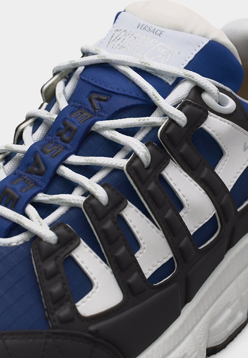 Baskets Versace bleu et blanc avec accents noirs, lacets blancs, et "Versace" embossé sur la languette et la protection des lacets.