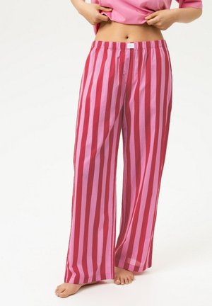 Personne portant un pantalon de pyjama ample à rayures verticales roses et rouges, soulevant un haut rose assorti, pieds nus sur fond blanc.