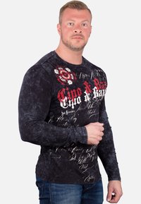 Langarmhemd in Schwarz mit strukturiertem Muster, mit roten und weißen Grafikdrucken und Schriftzügen auf der Vorderseite, kombiniert mit blauen Jeans.