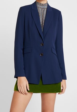 Kobieta w granatowej, dopasowanej marynarce na dwa guziki, wzorzystej bluzce z wysokim kołnierzem i krótkiej oliwkowozielonej spódnicy.