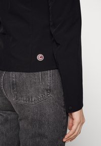 Černá bunda s hladkou texturou, se zaobleným logem v bordó barvě a rukávem na knoflíky. Nosená přes šedé džíny.