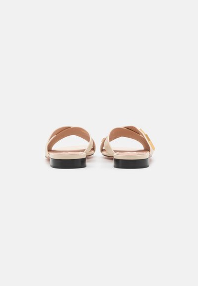 Flat sandals with a beige leather upper, featuring crisscross straps and a black rubber sole. Minimalistic design with a metallic accent.