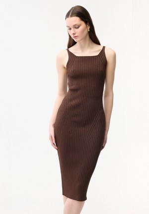 Kobieta w brązowej, prążkowanej sukience midi bez rękawów z kwadratowym dekoltem i złotymi kolczykami-kołami, stojąca na białym tle.