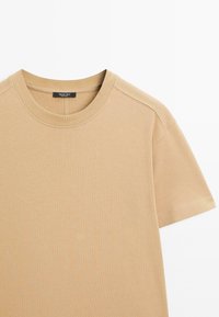 T-shirt in cotone beige con collo rotondo, maniche corte e finitura pulita. Presenta una leggera texture e un'etichetta con logo sul collo.
