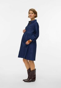 Tamno plava haljina s nabranym strukom i dugmadi na izrezu, s dugim rukavima. Upućena uz tamno smeđe kaubojke.