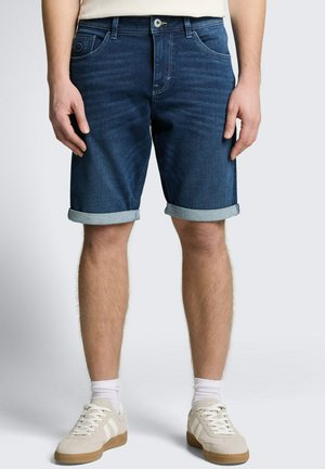 Homme portant un short en jean bleu foncé retroussé, des chaussettes blanches et des baskets beiges, debout les bras détendus le long du corps.