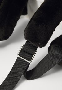 Pelliccia sintetica nera con una texture morbida, presenta una tracolla nera regolabile con una fibbia argentata, parzialmente visibile su uno sfondo bianco.