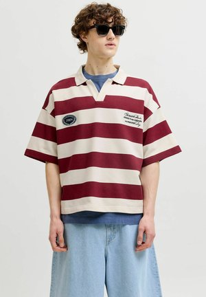 Junge Person trägt schwarze Sonnenbrille, rugby Shirt mit bordeauxroten und weißen Streifen und Applikationen, blaues Unterhemd und hellblaue Jeans mit weitem Bein.
