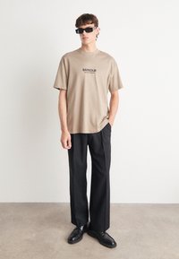 Beige puuvillane t-särk lühikeste varrukatega, millel on must trükk "Barbour International". Koos kõrge vöökohtadega mustade pükste ja mustade kingadega.