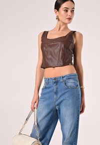 Top corto in pelle marrone con scollatura quadrata, abbinato a jeans a vita alta blu chiaro. La modella tiene una borsa bianca con accenti argentati.