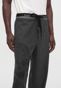 Homme portant un pantalon gris foncé à fines rayures avec un motif subtil floral et serpent noir, et une ceinture noire à cordon avec texte blanc.