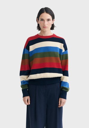 Femme aux cheveux courts et foncés portant un pull rayé multicolore et un pantalon large foncé sur un fond clair uni.
